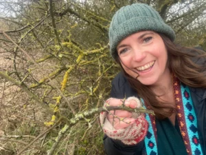 Smiling woman wearing a beanie holding a twig with branches of a tree behind her.